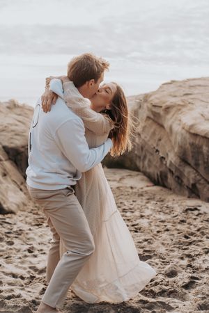 Couple shares intimate moment on sandy beach with coastal rocks in background.