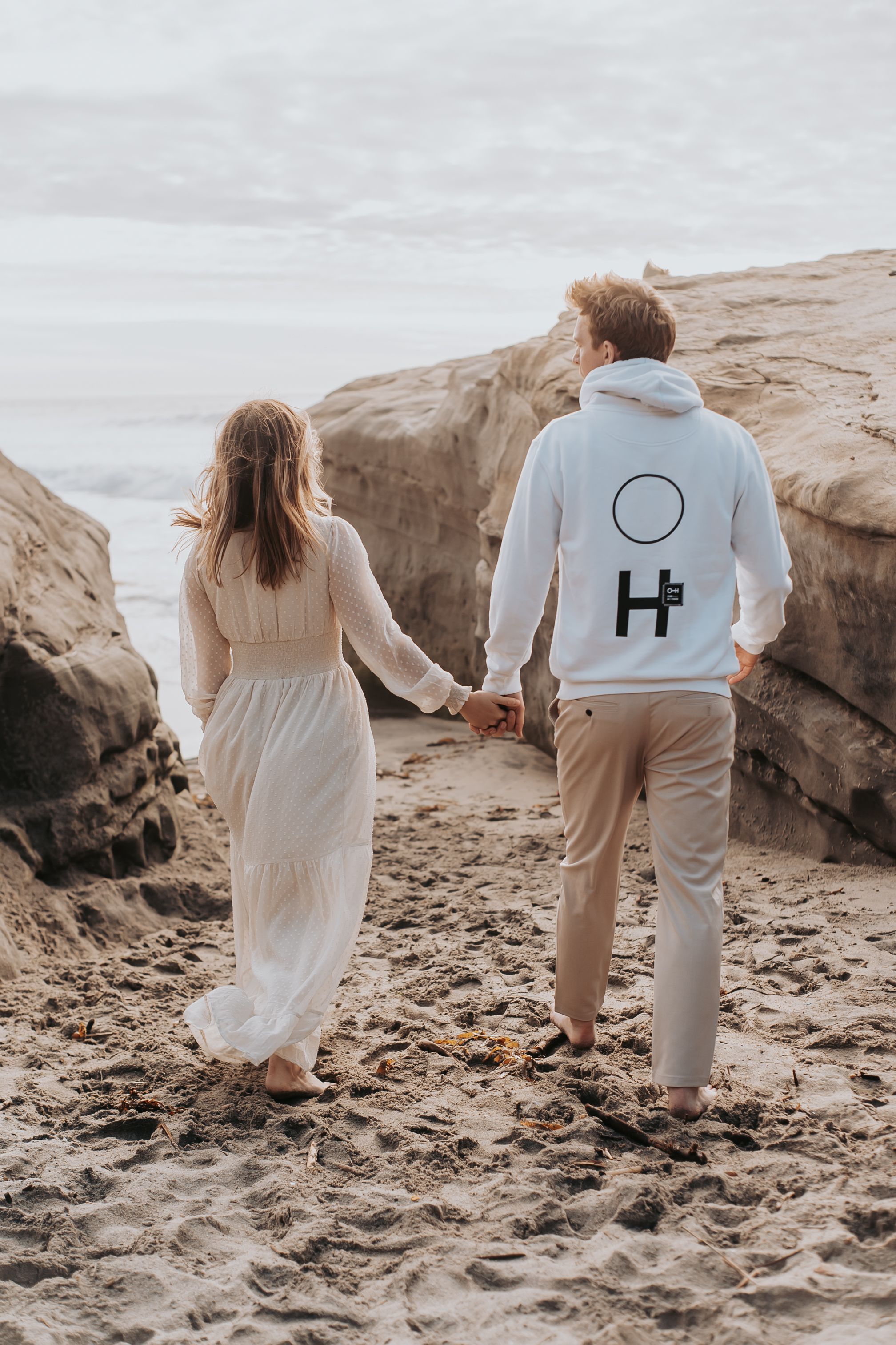 Couple walks hand in hand on rocky beach wearing matching white and beige outfits.