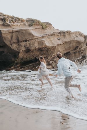 Couple splashes through ocean waves near rocky cliffs at beach.