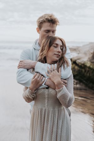Intimate coastal portrait of couple embracing in muted tones by rocky beach.