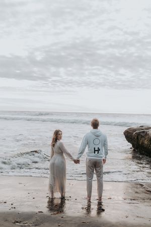 A romantic couple stands hand in hand at the ocean's edge during sunset, waves lapping at their feet.