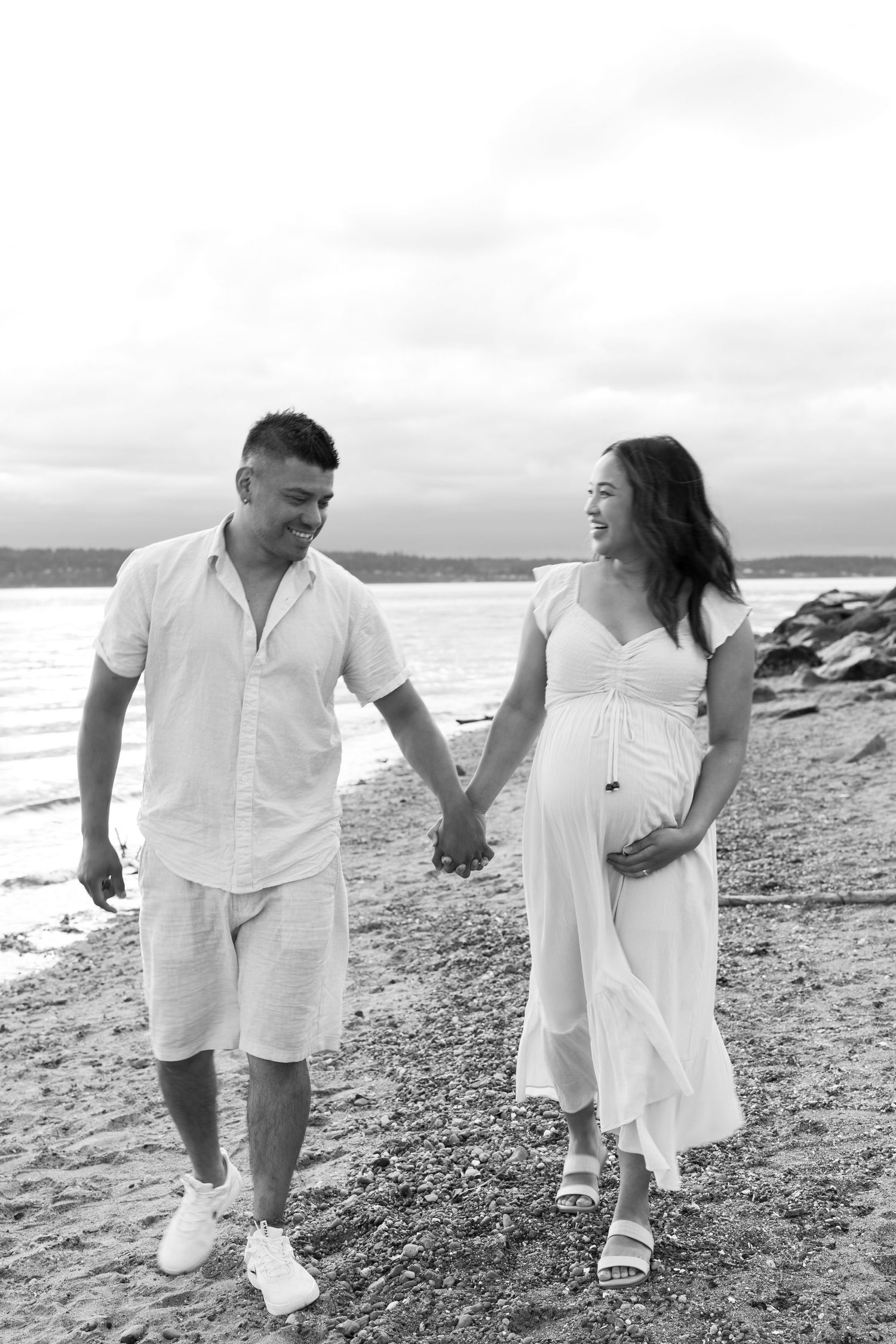 A couple walks hand in hand along a rocky beach in black and white.