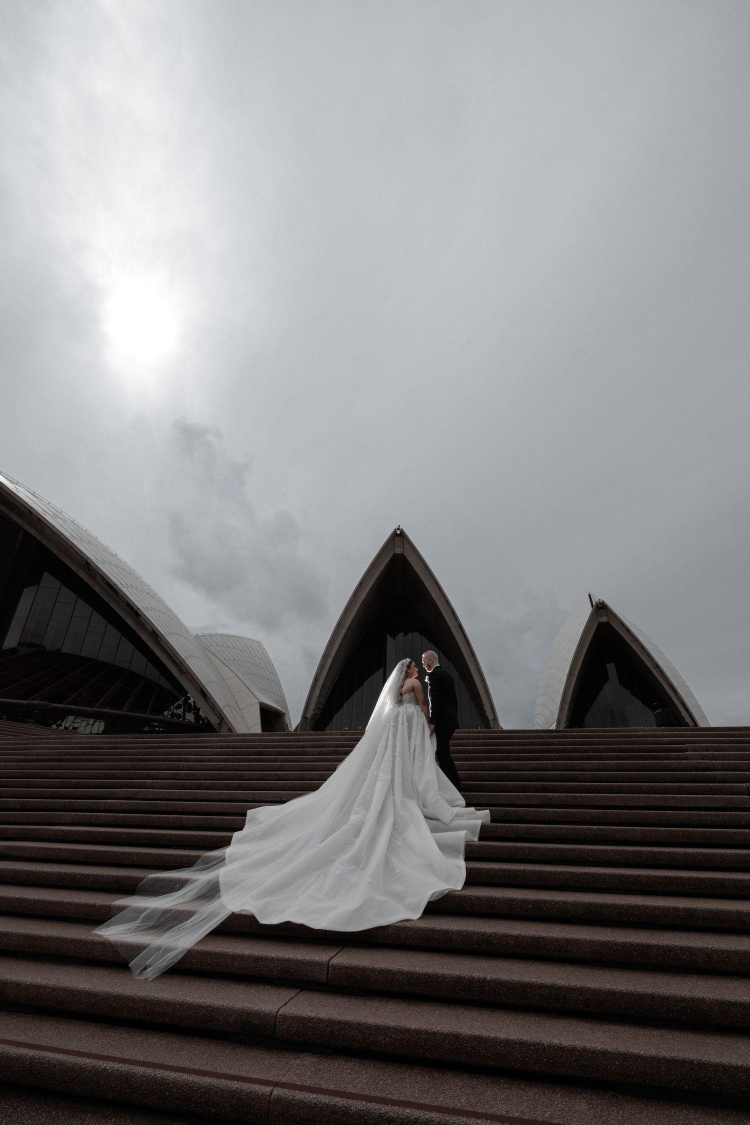 Bride in flowing white gown and veil stands on steps of Sydney Opera House under cloudy sky.