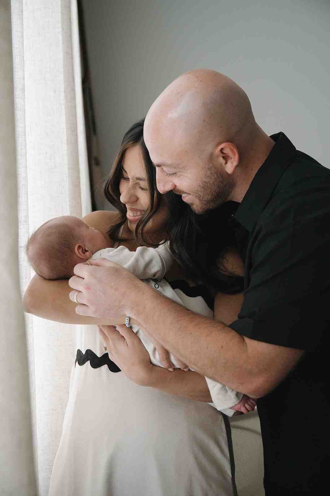 A couple shares a tender moment with their newborn baby near a bright window in an intimate indoor setting.