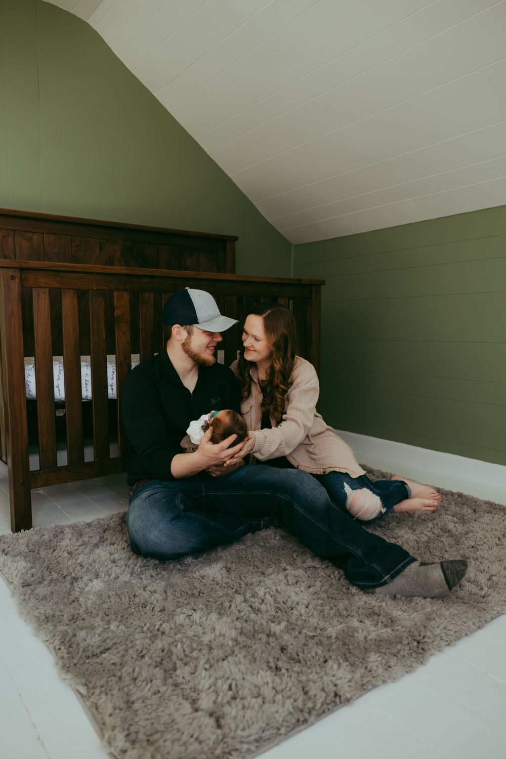 Intimate family photo in nursery with parents cradling newborn on plush gray carpet.