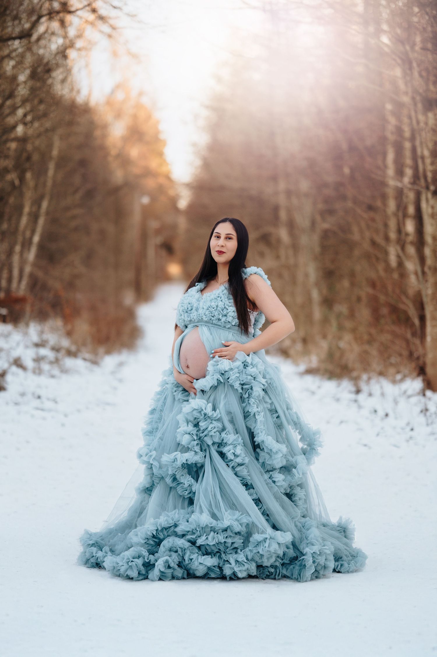 Ethereal maternity portrait in flowing blue gown against snowy winter forest backdrop.