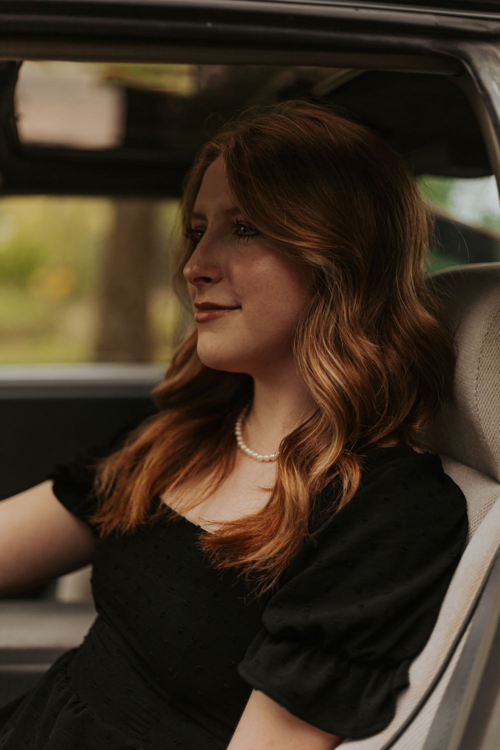 Woman with wavy hair wearing pearl necklace sitting in car driver's seat.