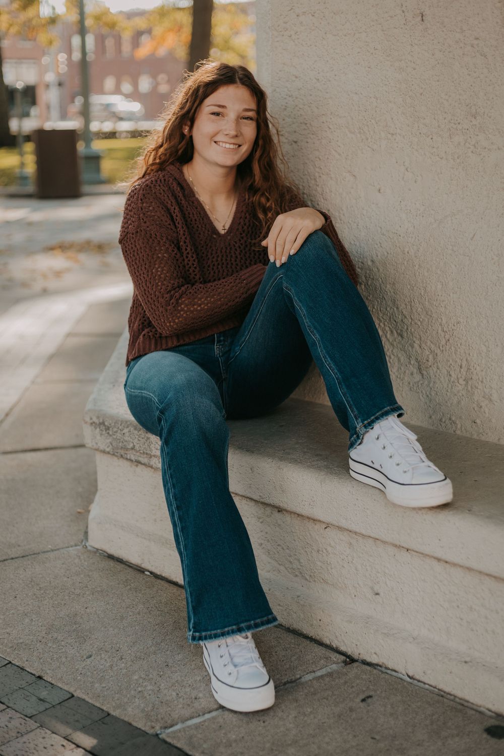Casual style featuring brown sweater, jeans and white sneakers on concrete steps.
