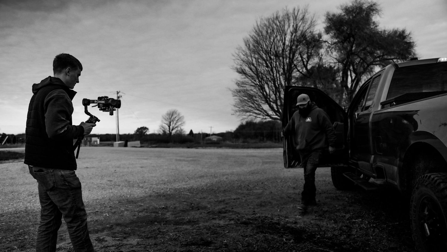 Filmmaker using gimbal stabilizer to film a man standing by a pickup truck in an open field.