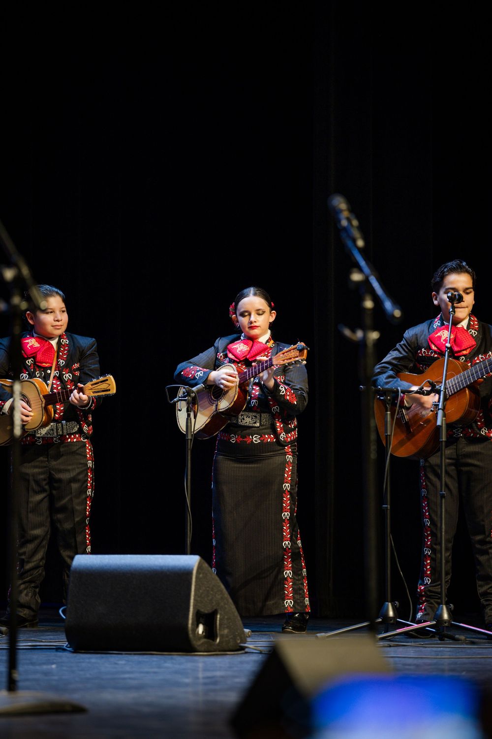 A mariachi musician performs on acoustic guitar while wearing traditional black attire with embroidered trim on stage.