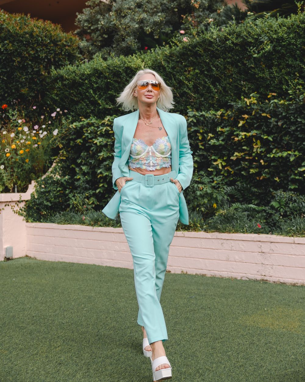 Fashion model in mint green suit poses playfully on grass against garden backdrop.