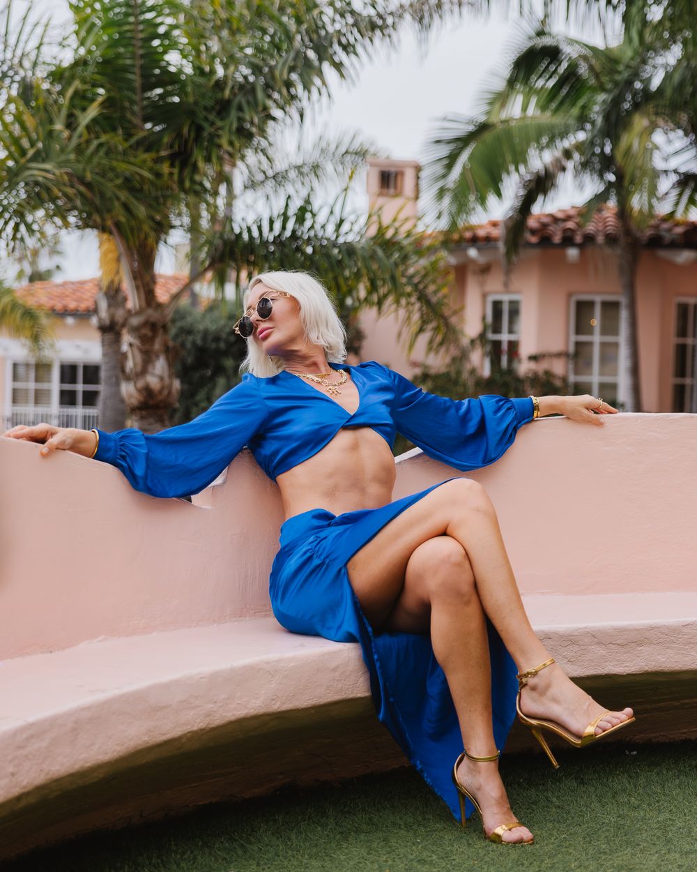 Model reclines dramatically along pink wall with tropical landscaping.