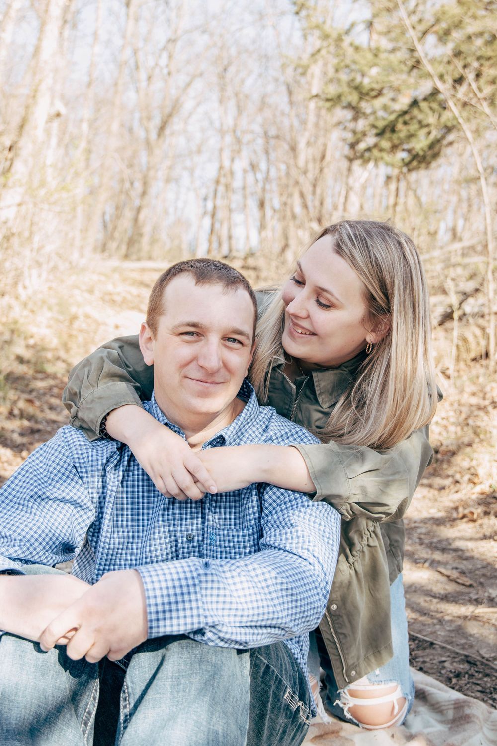 A couple shares a tender moment while sitting on a blanket in a sunlit forest setting.
