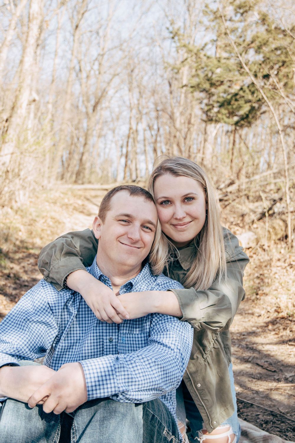 A couple poses for a casual portrait while sitting outdoors on a wooded trail.