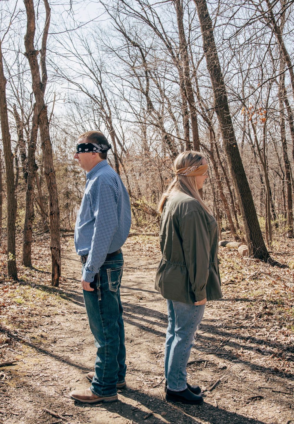 A couple stands back-to-back on a forest trail surrounded by bare trees on a sunny day.