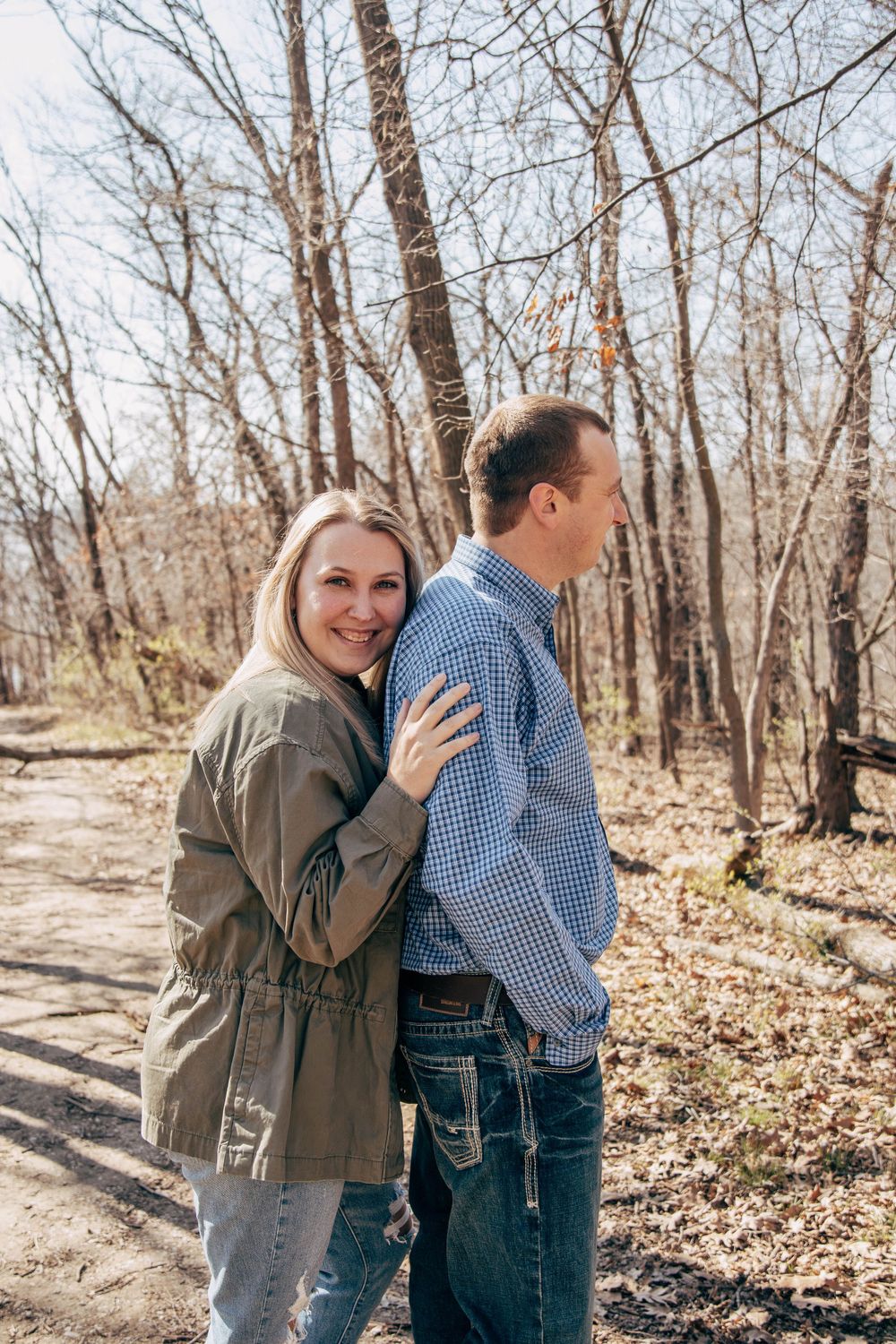 A couple embracing on a forest path surrounded by bare trees and fallen leaves.