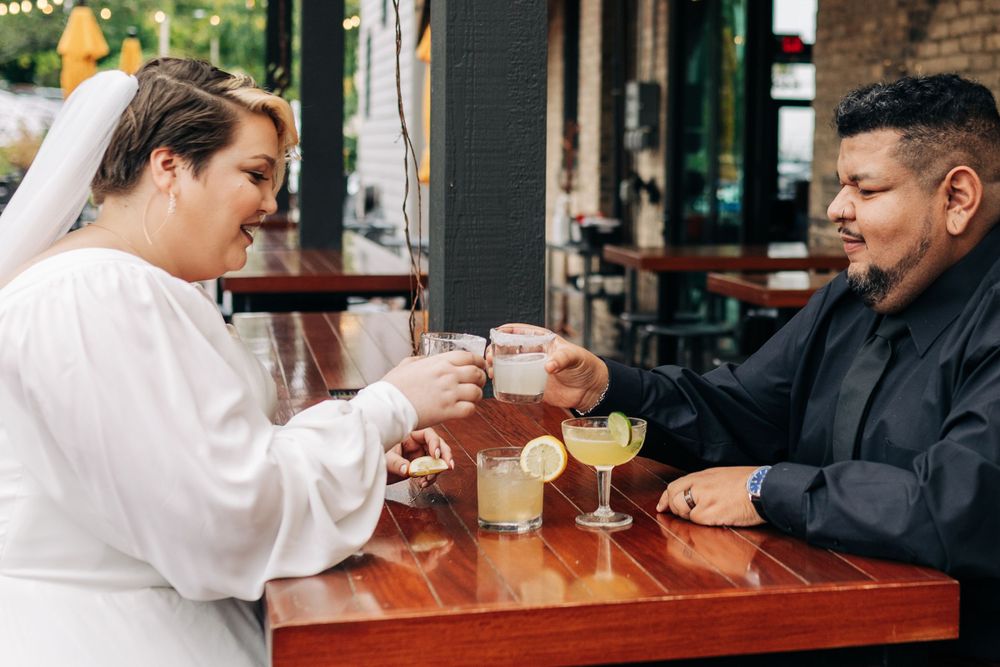A couple toasts with cocktails at a wooden table in a restaurant.