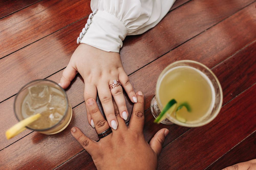 Two hands with wedding rings intertwined next to cocktails on a wooden table.