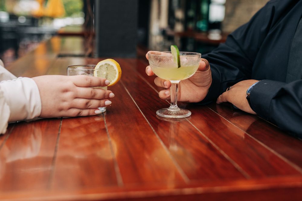 Close-up of hands holding citrus cocktails across a wooden table.