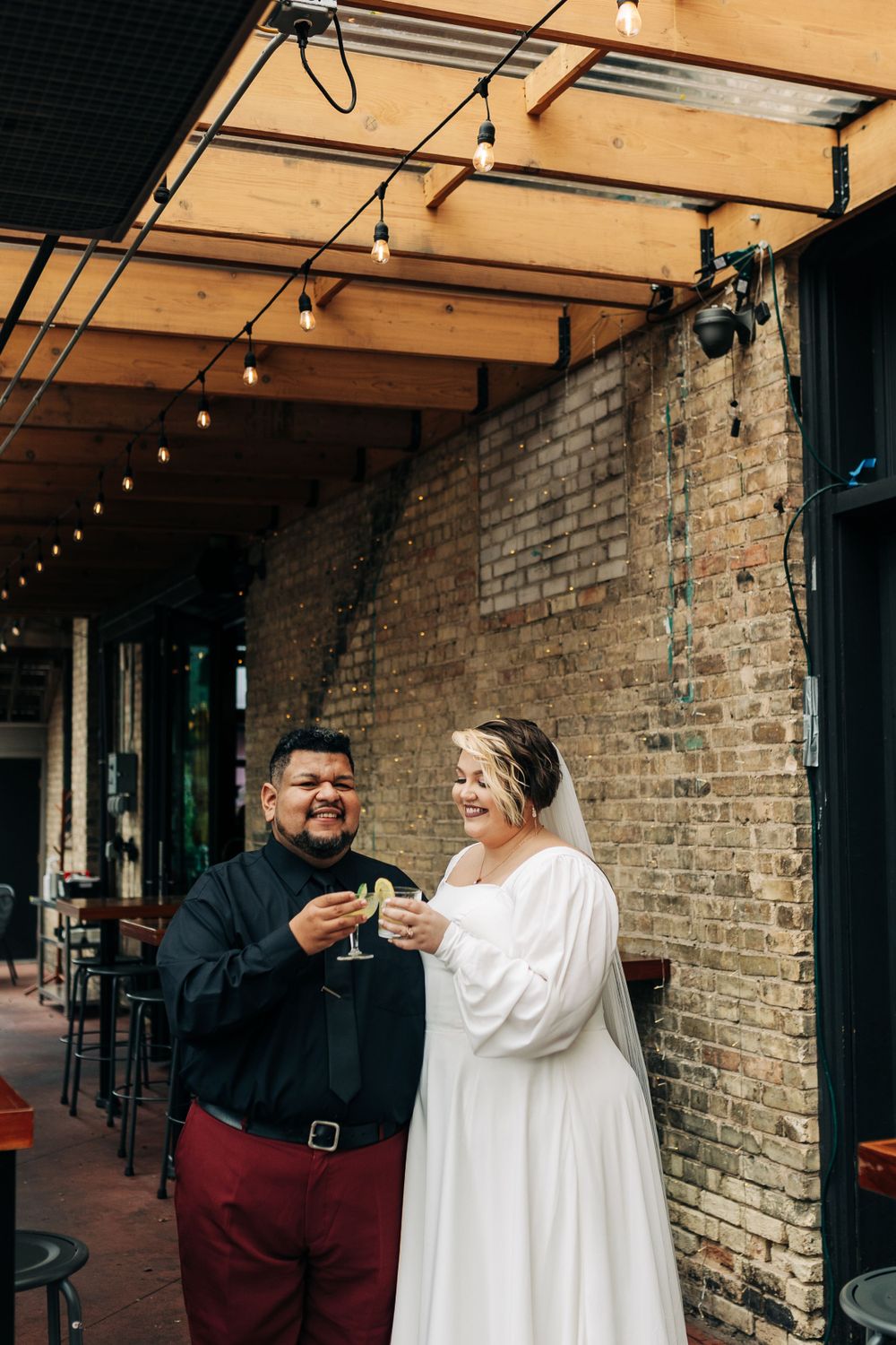 A couple toasts with margaritas against an exposed brick wall with string lights.