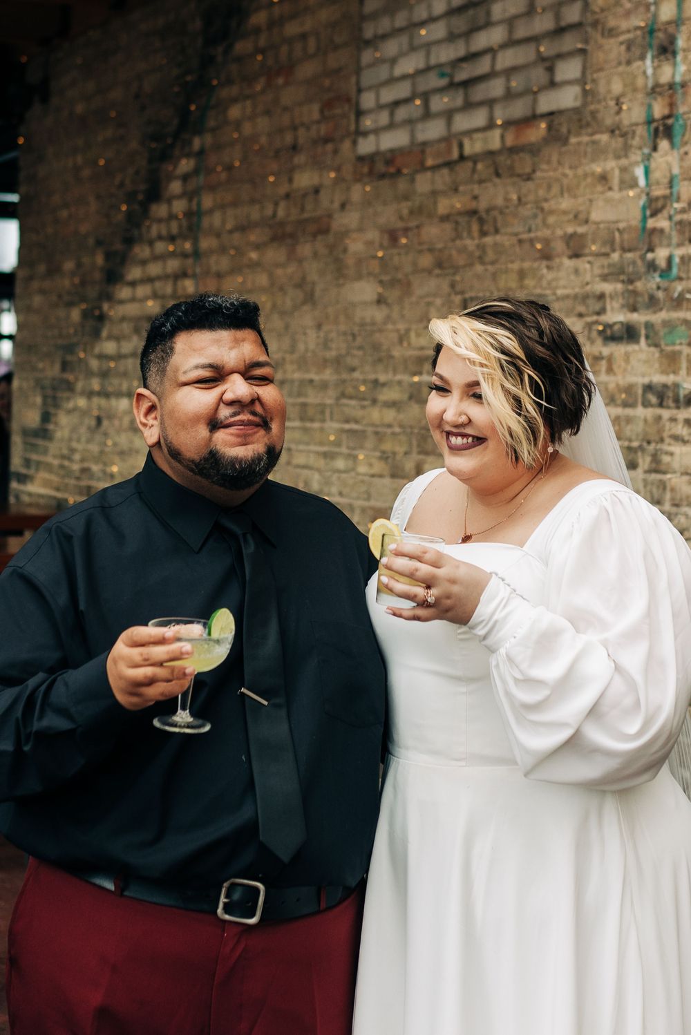 A couple enjoying cocktails and laughter against a rustic brick wall backdrop.