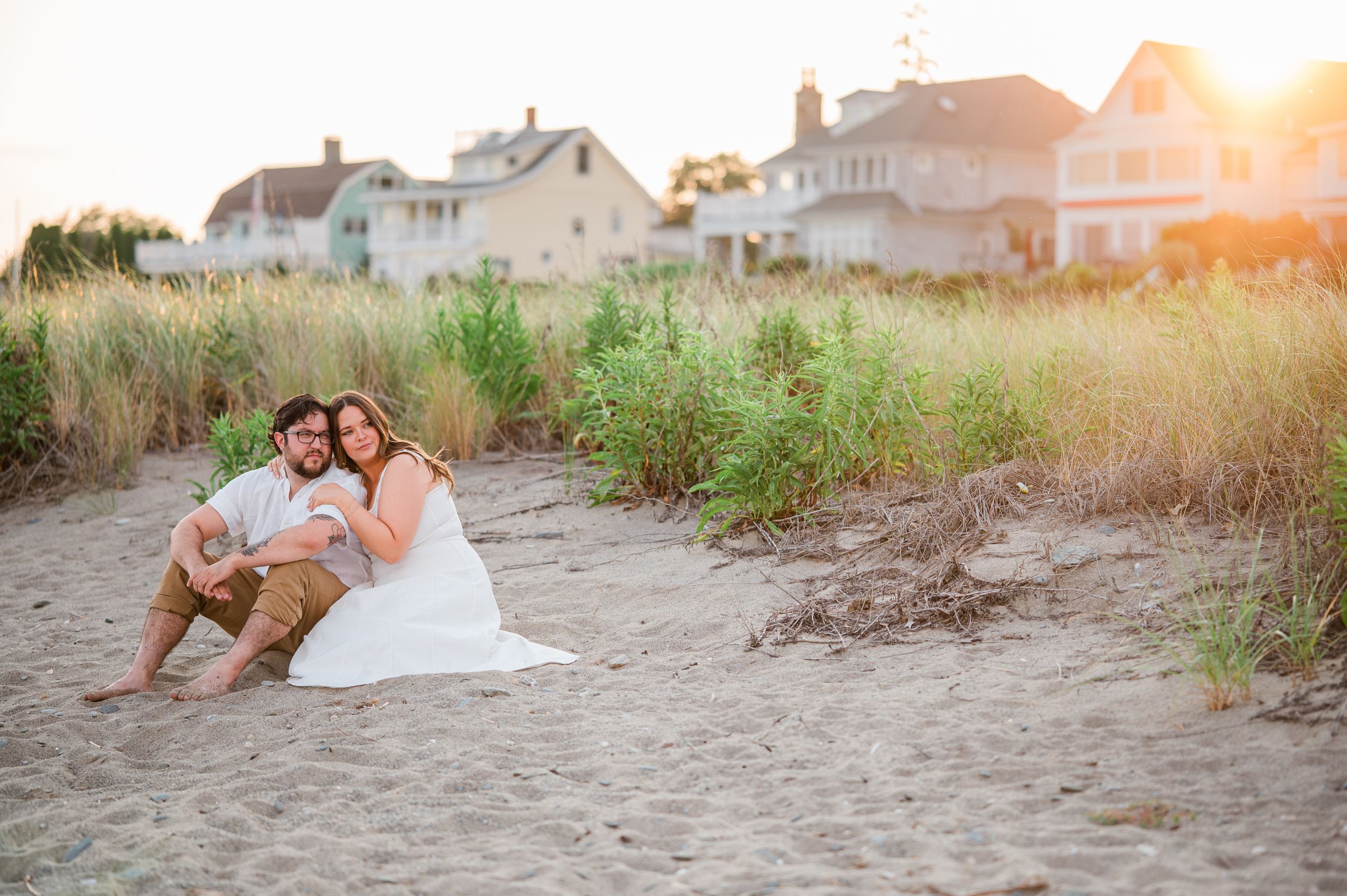 A couple sits together on a sandy beach at sunset with coastal homes in the background at the wall in Stratford, CT.