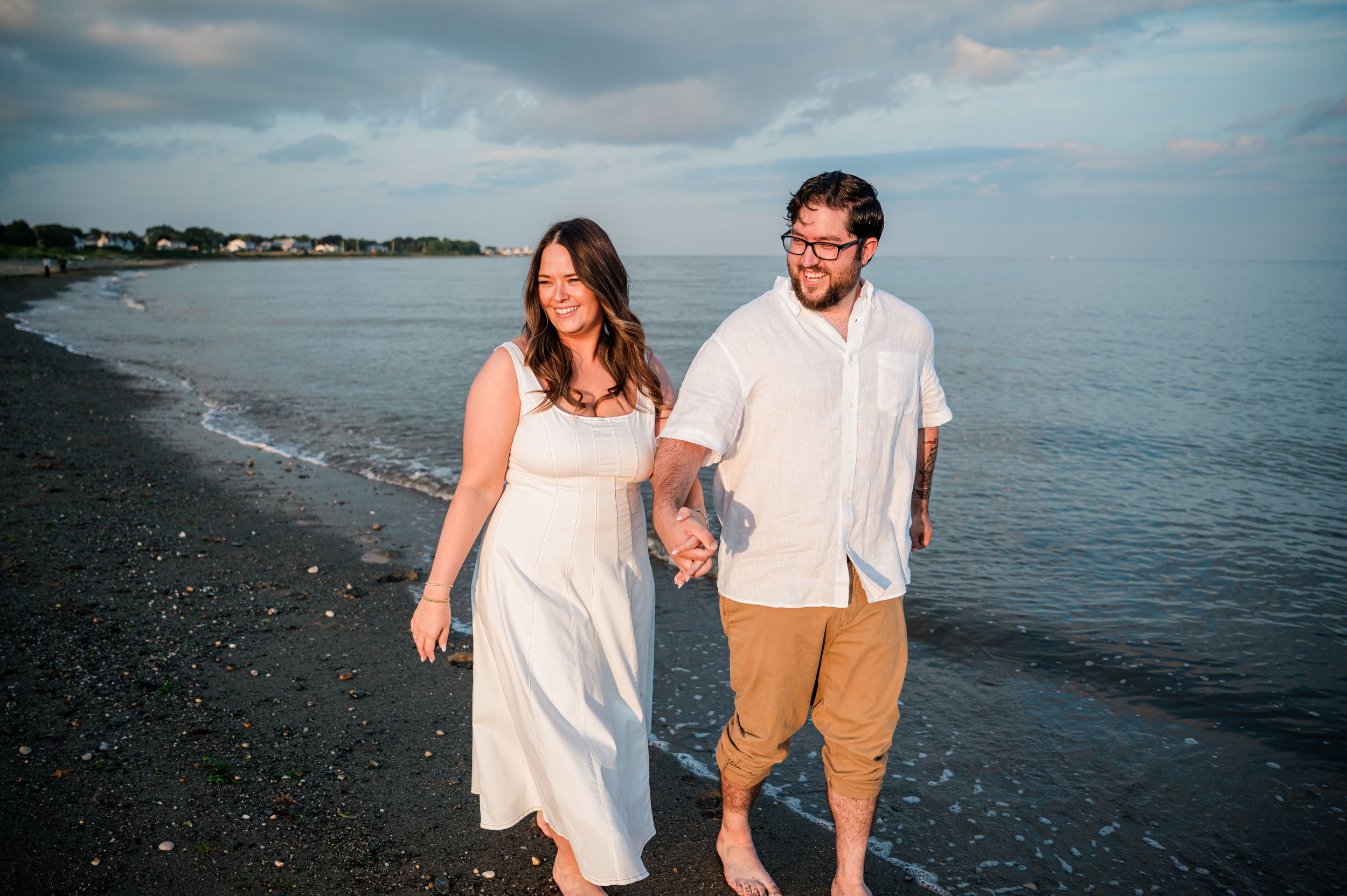 A couple walks hand in hand along a beach at sunset with the ocean and cloudy sky in the background at the wall in Stratford, CT.