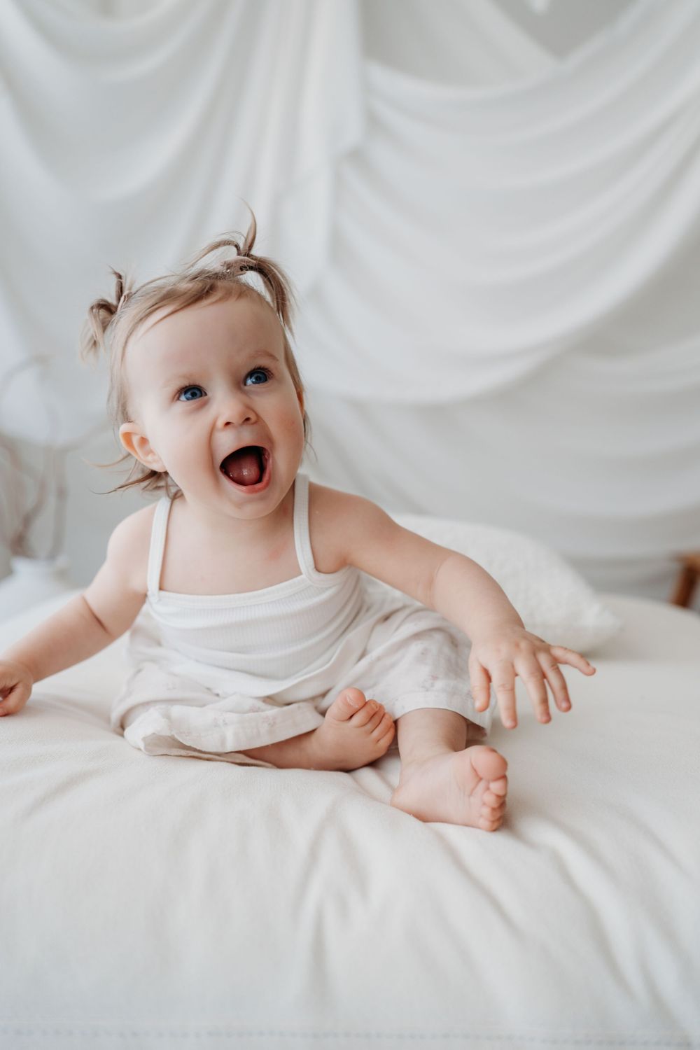 An adorable blue-eyed baby girl in white outfit sits in studio.