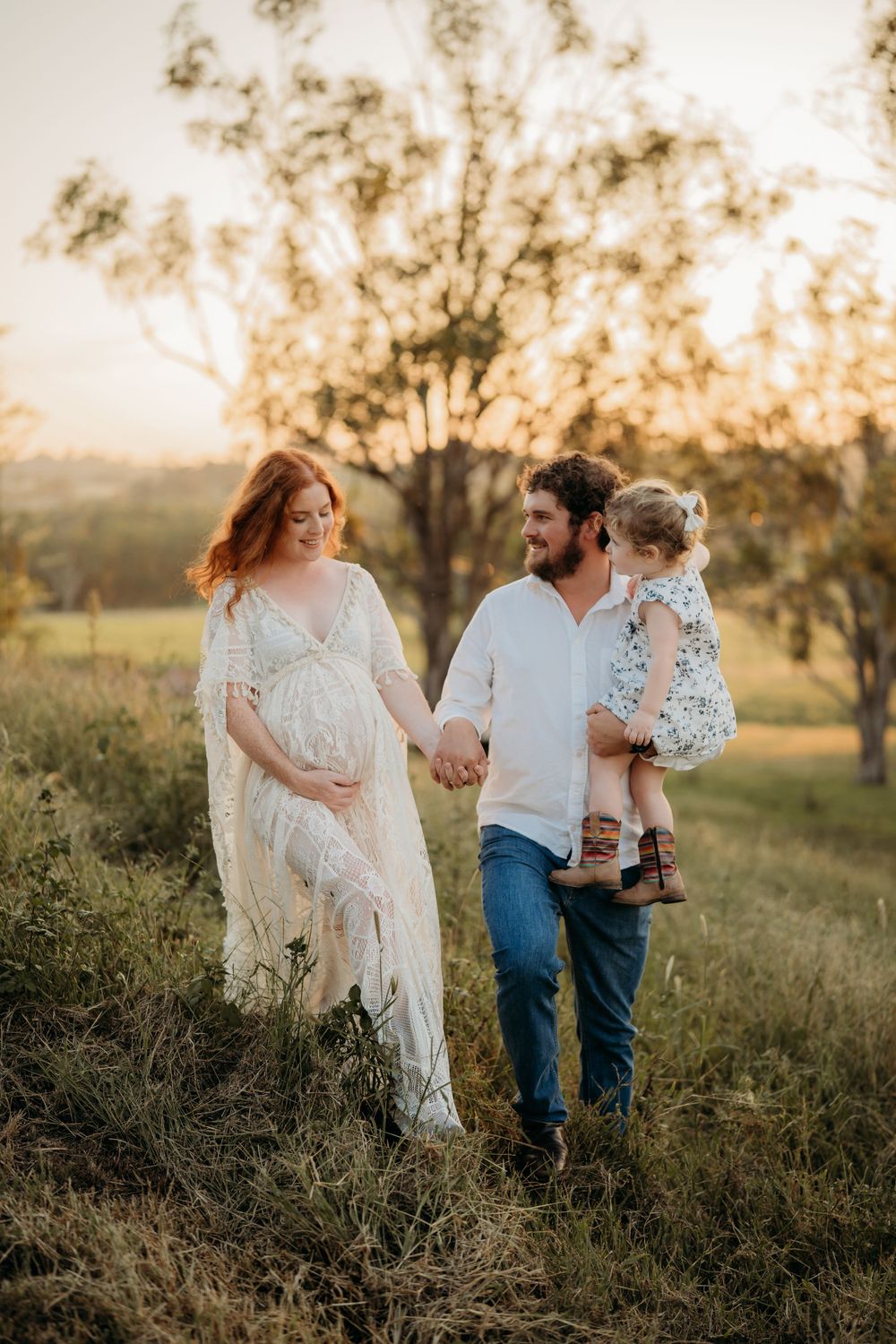 Expecting family walks hand-in-hand through grassy field at sunset, father carrying young child.