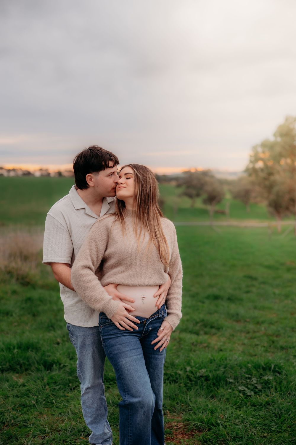 A couple shares a tender kiss in a green field at sunset, with the man gently holding the woman's baby bump.