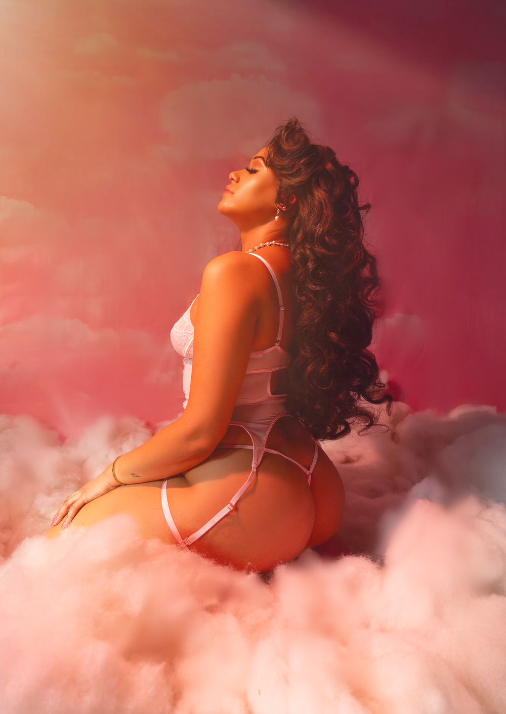Dreamy boudoir photoshoot with model posing on pink cloudy background.