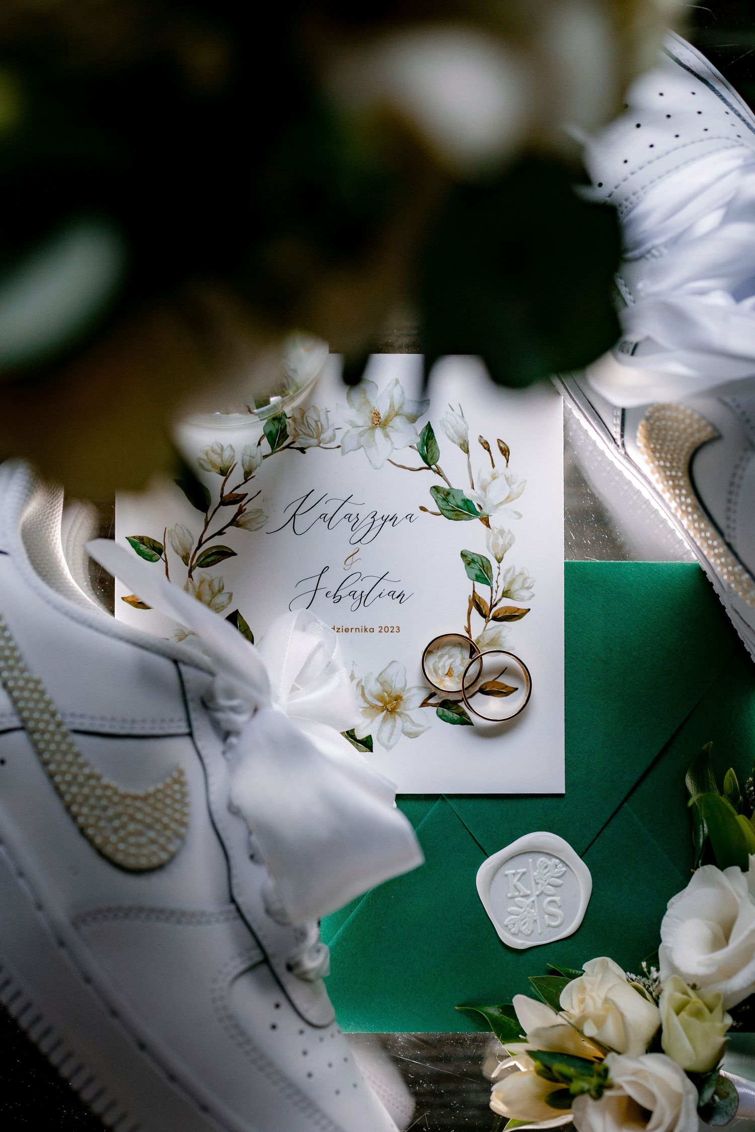 Wedding details flat lay with rings, white shoes, invitation and floral elements on green background.