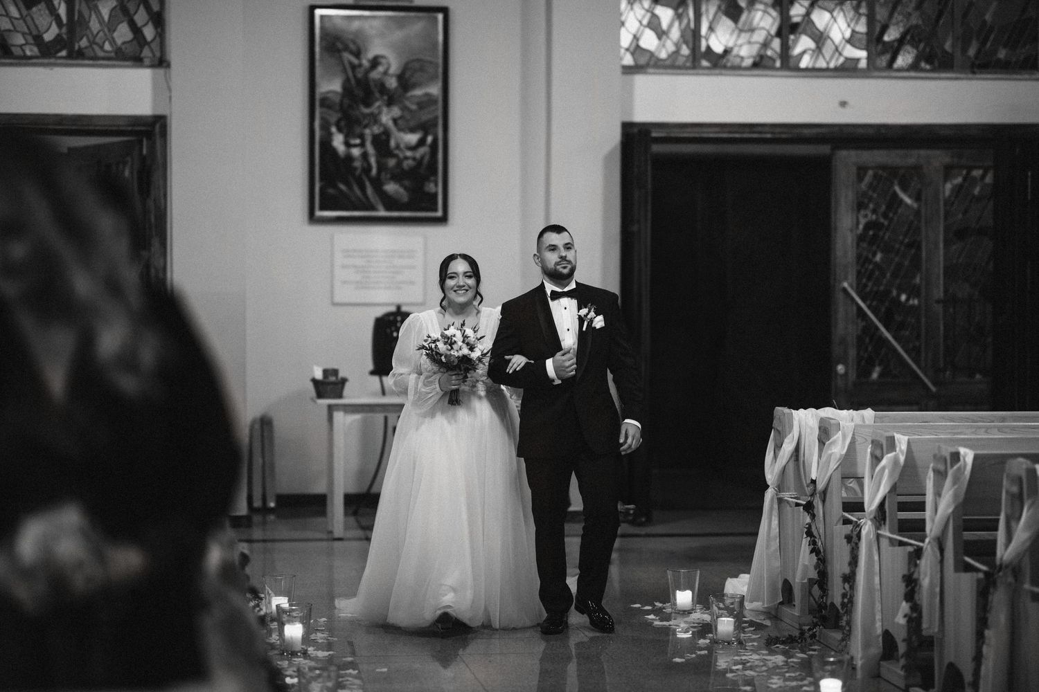 A black and white photo of a couple walking down the aisle decorated with candles.