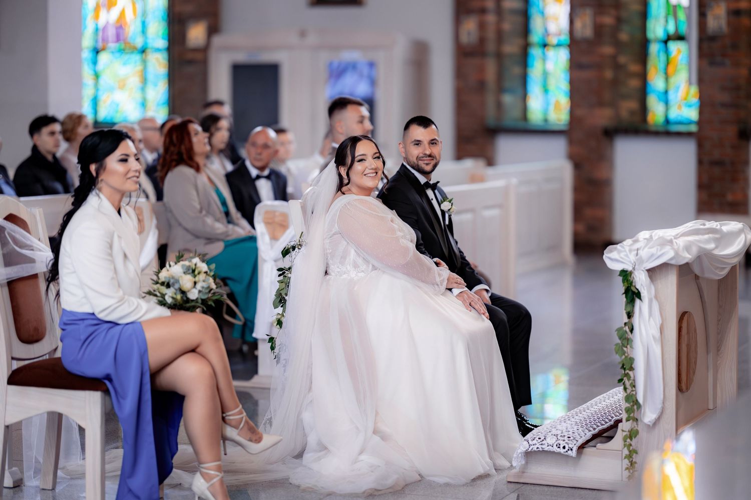 A wedding ceremony takes place in a church with stained glass windows and guests seated in rows.