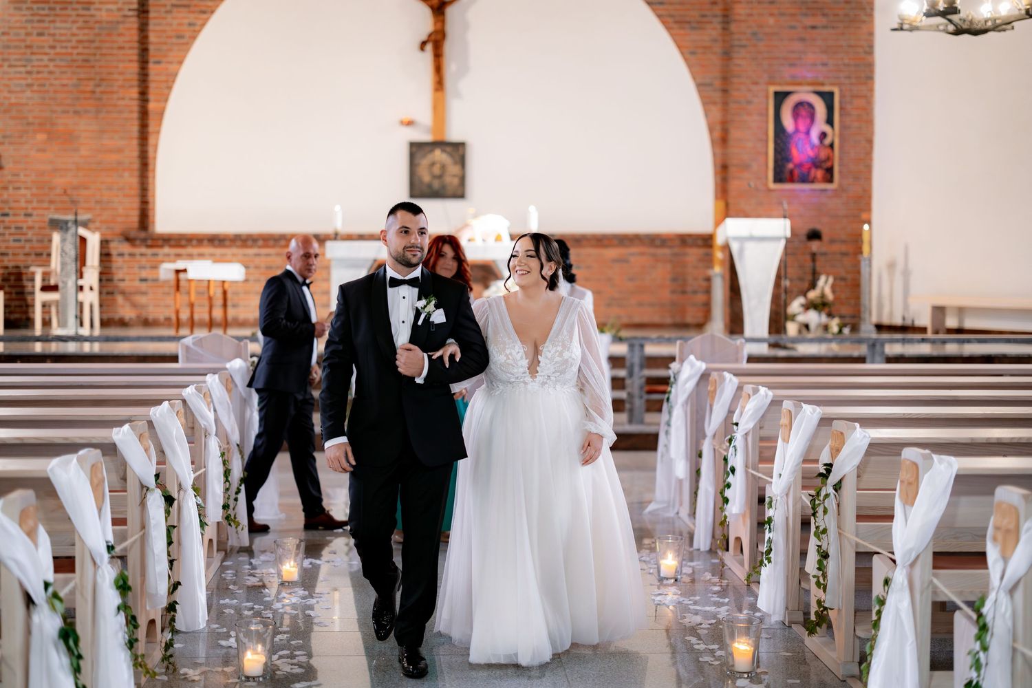 A couple walks down the church aisle decorated with floral arrangements and white ribbons.