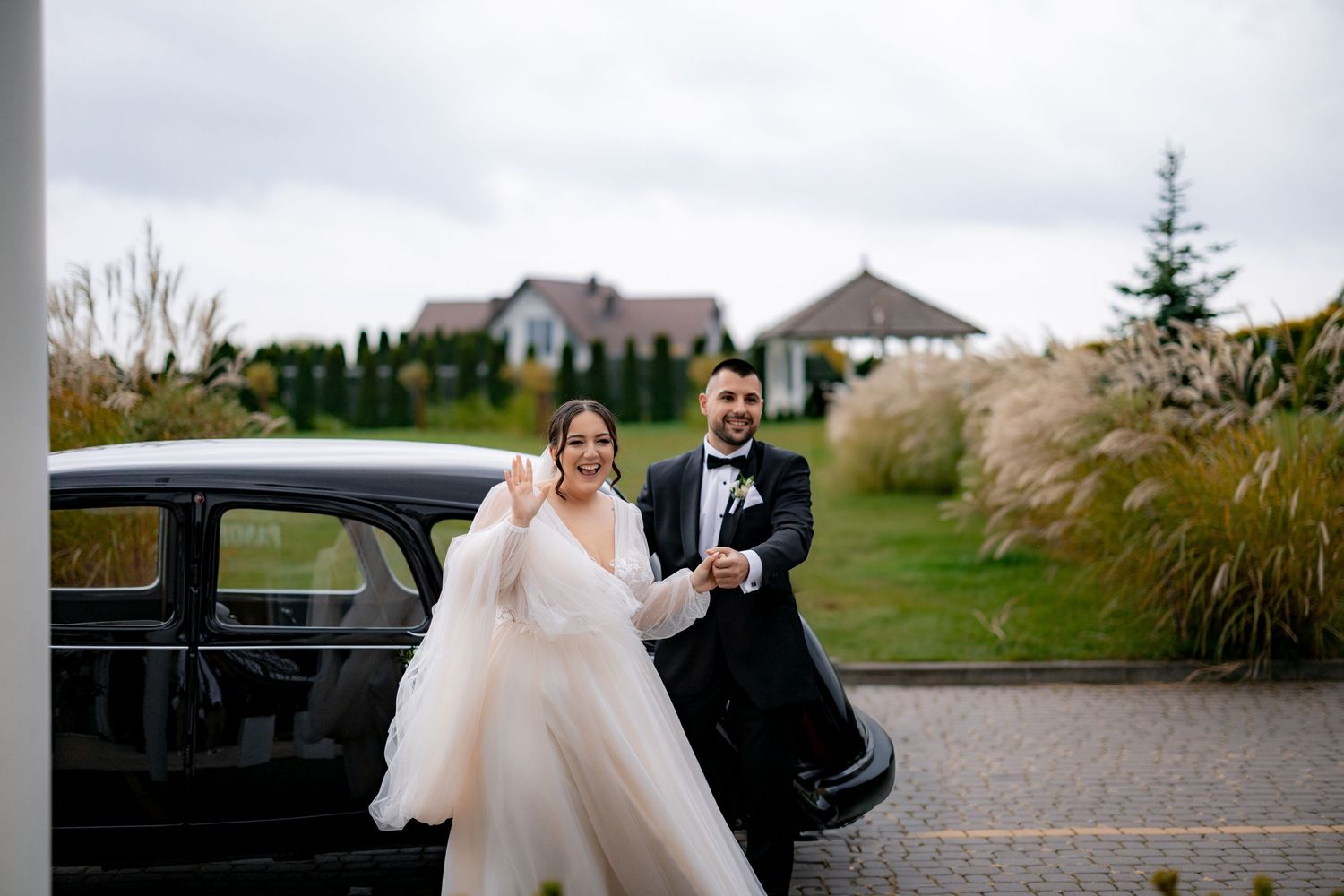 A couple poses next to a vintage black car with ornamental grasses in the background.