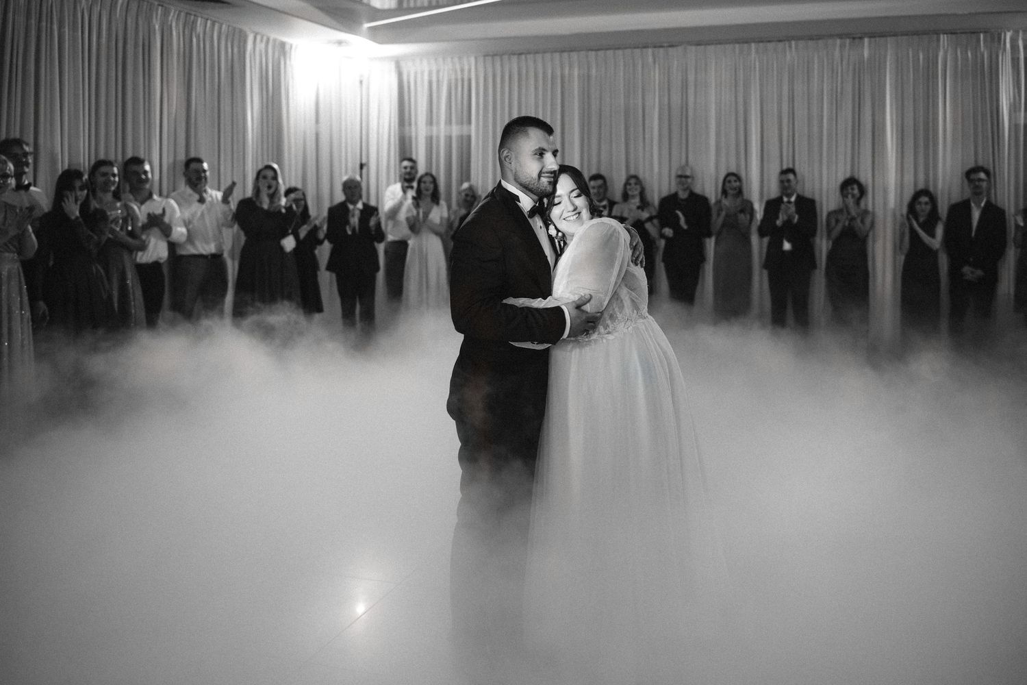 Wedding couple dances in a cloud of fog while guests watch from the edges of the dance floor.