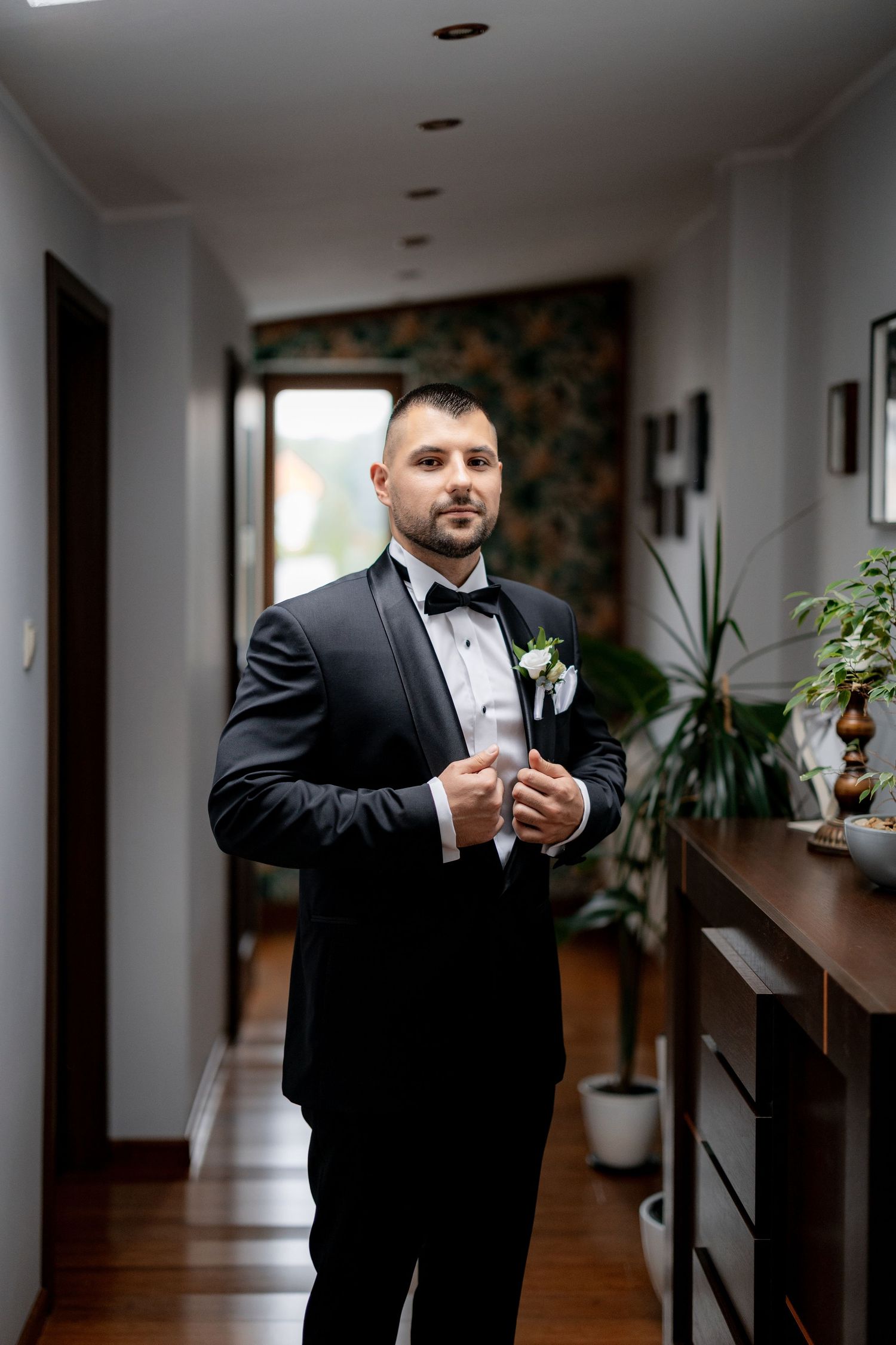 Groom in black tuxedo adjusts cufflinks while standing in hallway.