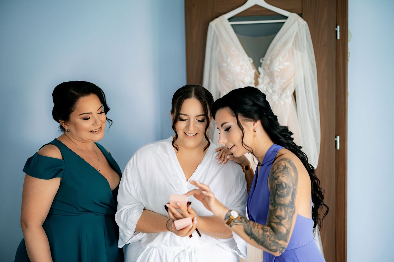 Three women share a tender moment while getting ready in a room with a white dress hanging in the background.