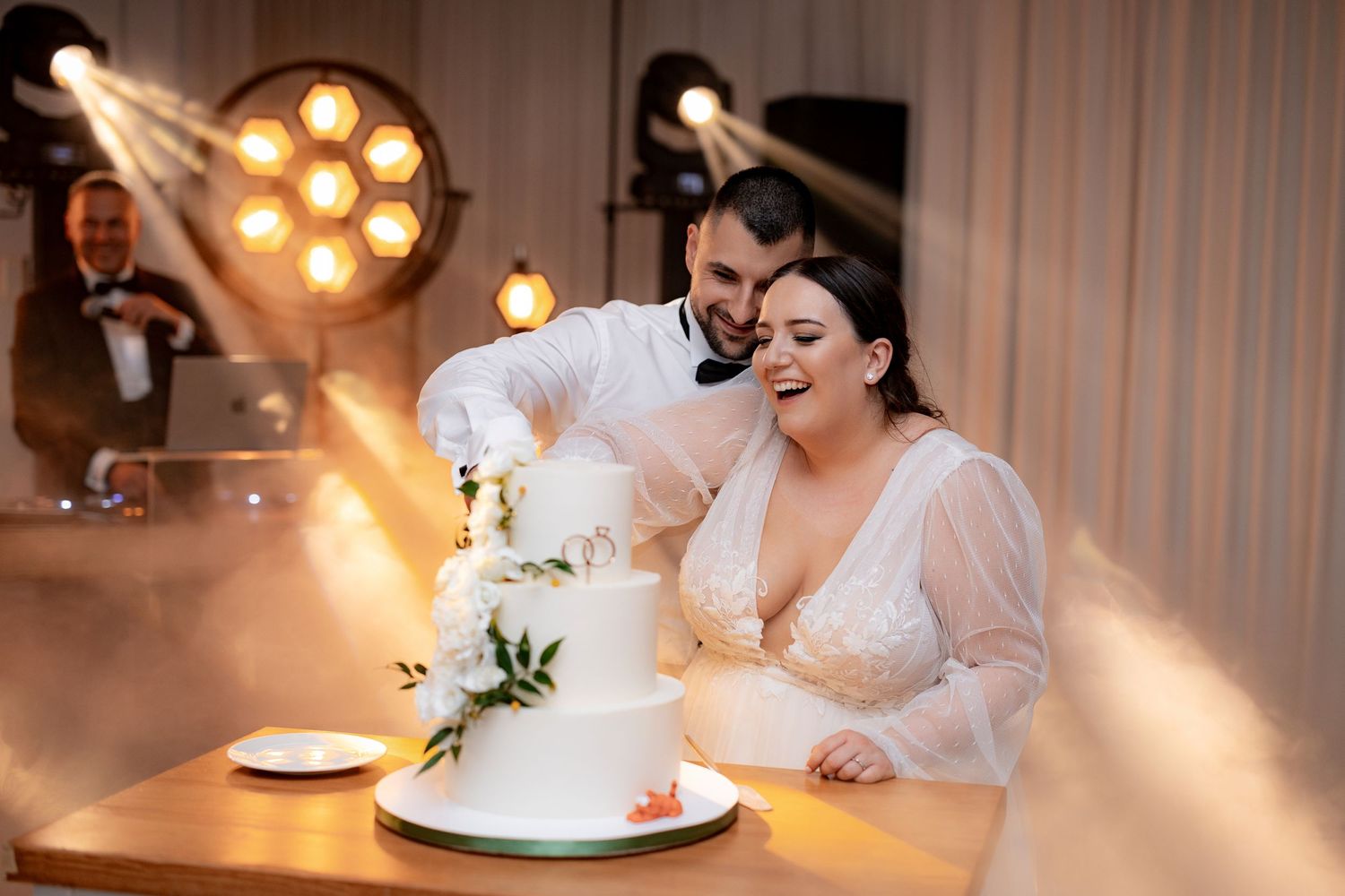 A couple cuts their elegant white wedding cake decorated with greenery in a warmly lit reception venue.