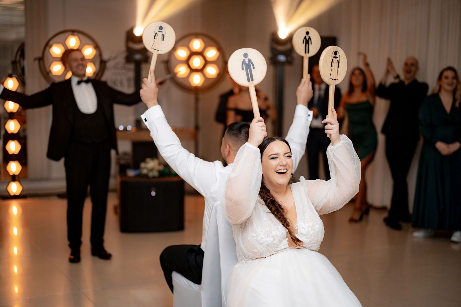 Wedding celebration with people dancing and holding up signs in a brightly lit reception hall.