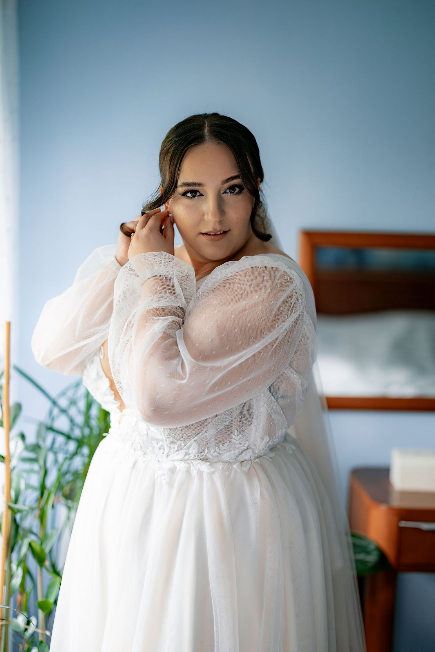 A beautiful plus size bride in a sheer white robe poses elegantly in a bedroom setting.
