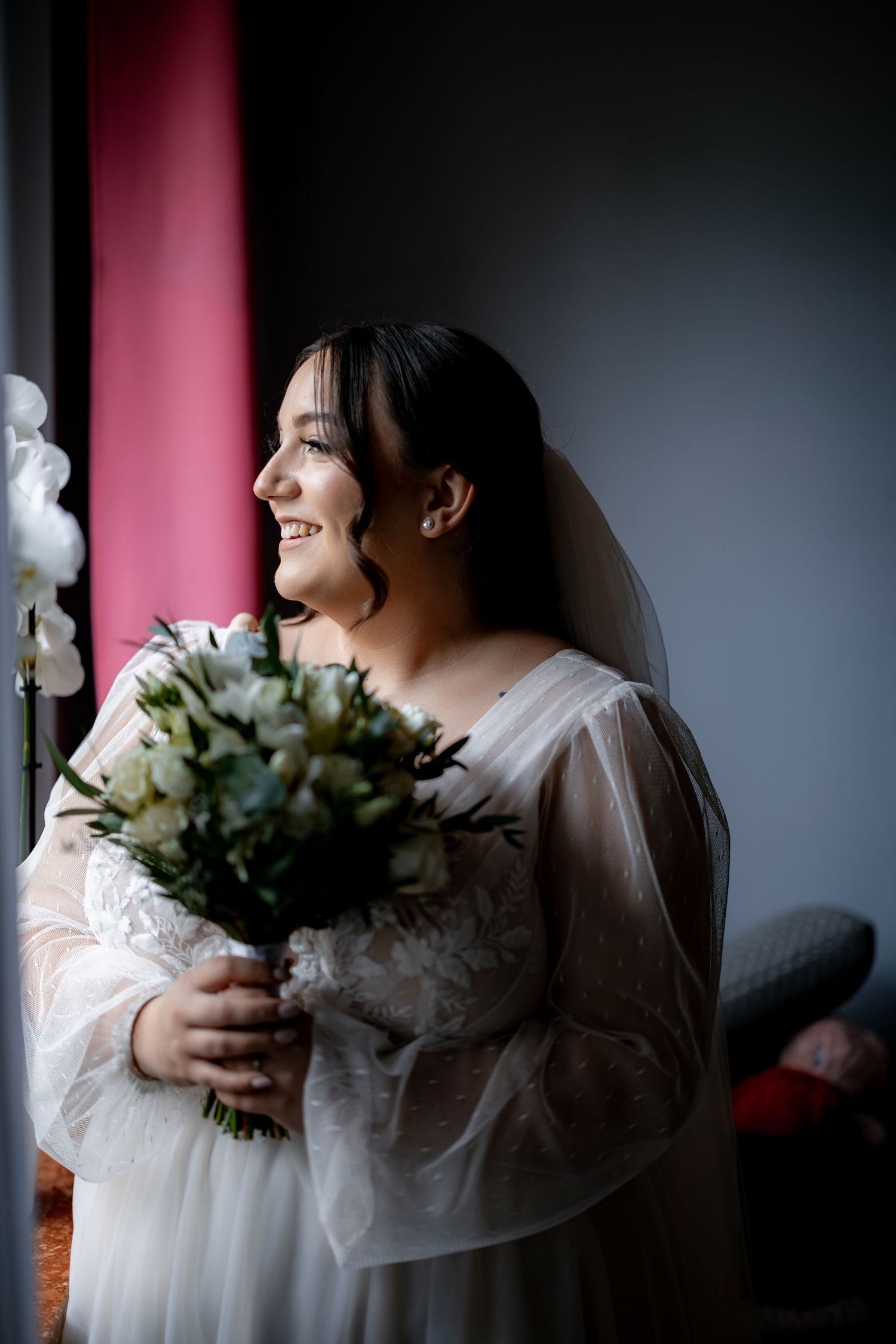 A radiant bride holds a white floral bouquet while smiling in natural window light.