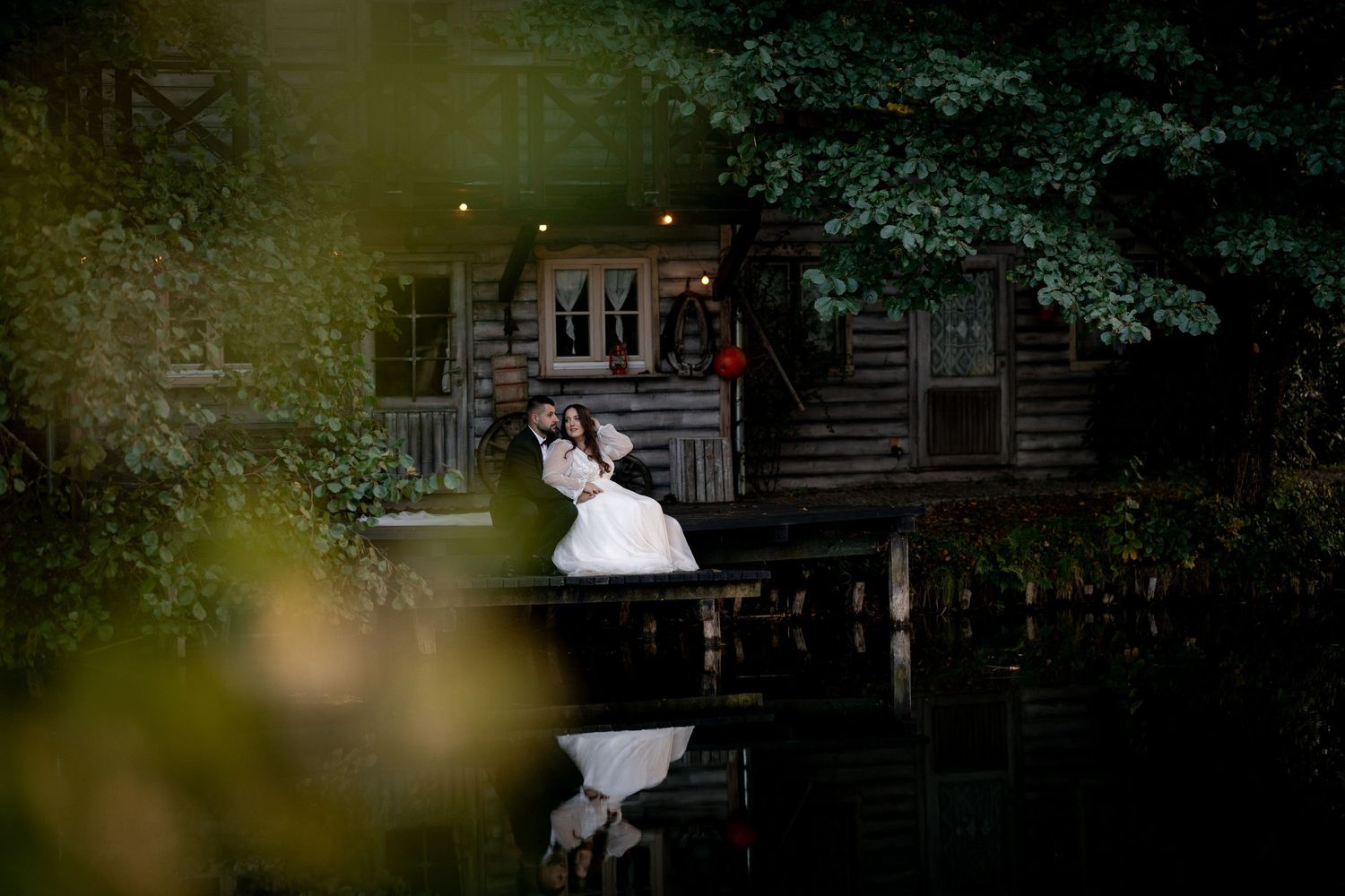 A dreamy wedding photo of a couple sitting on a dock by a rustic cabin at dusk.