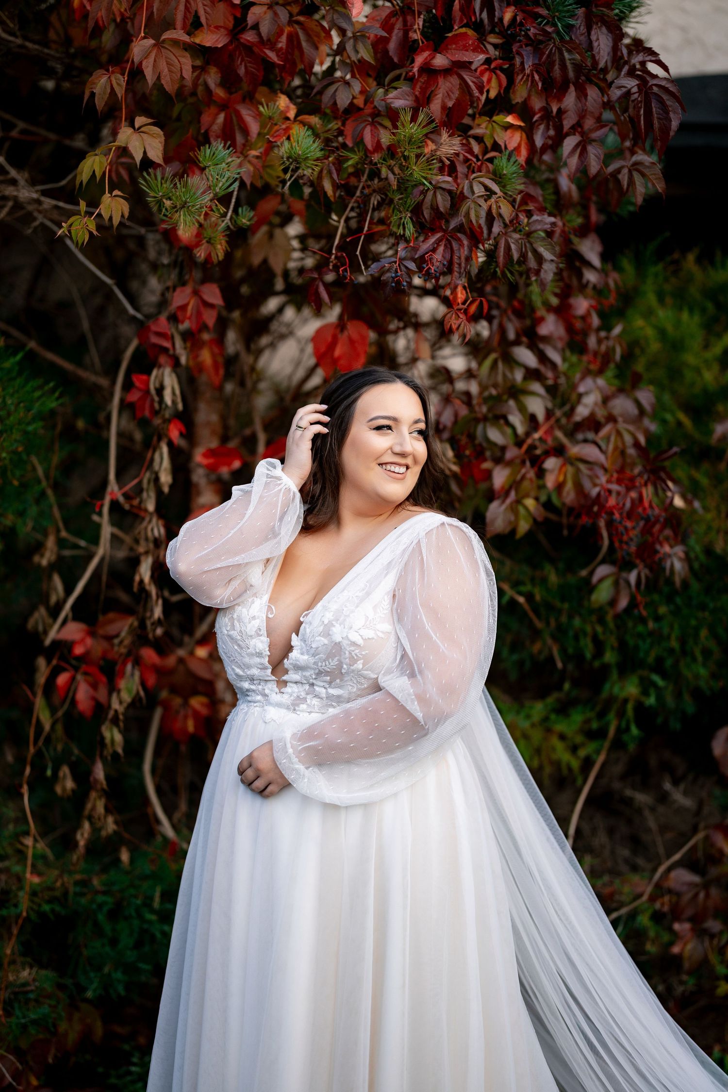 A bride in a flowing white dress poses in front of autumn foliage with red leaves.