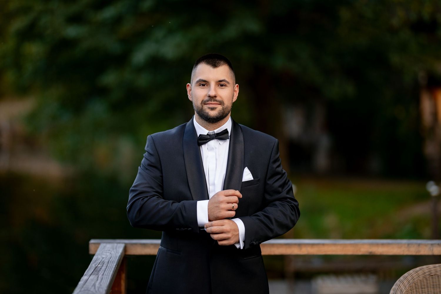 Groom in classic black tuxedo poses outdoors during golden hour.