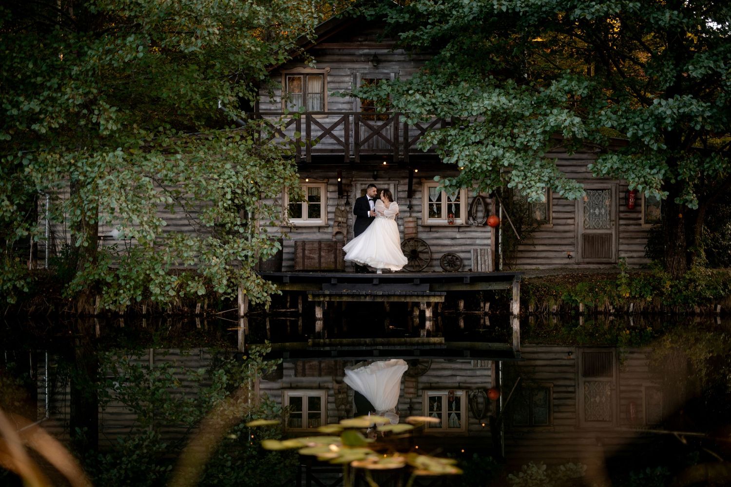 Wedding couple poses in front of rustic wooden house with reflection in pond.