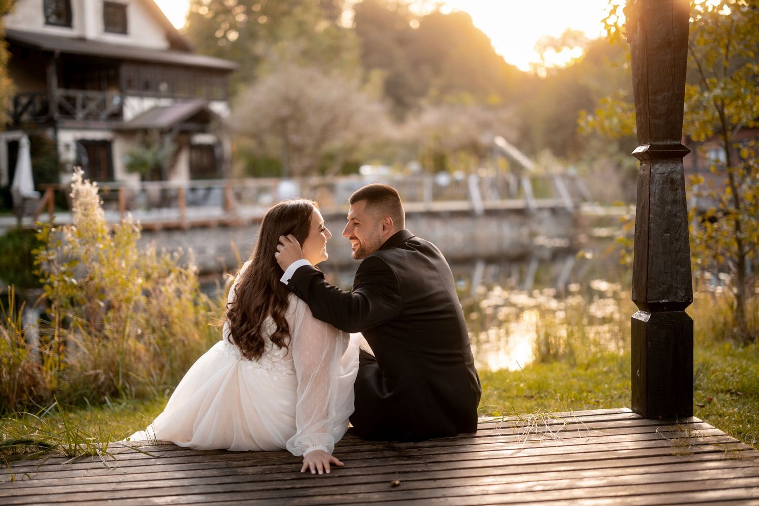 Couple sits on wooden dock at sunset with water and autumn scenery in background.