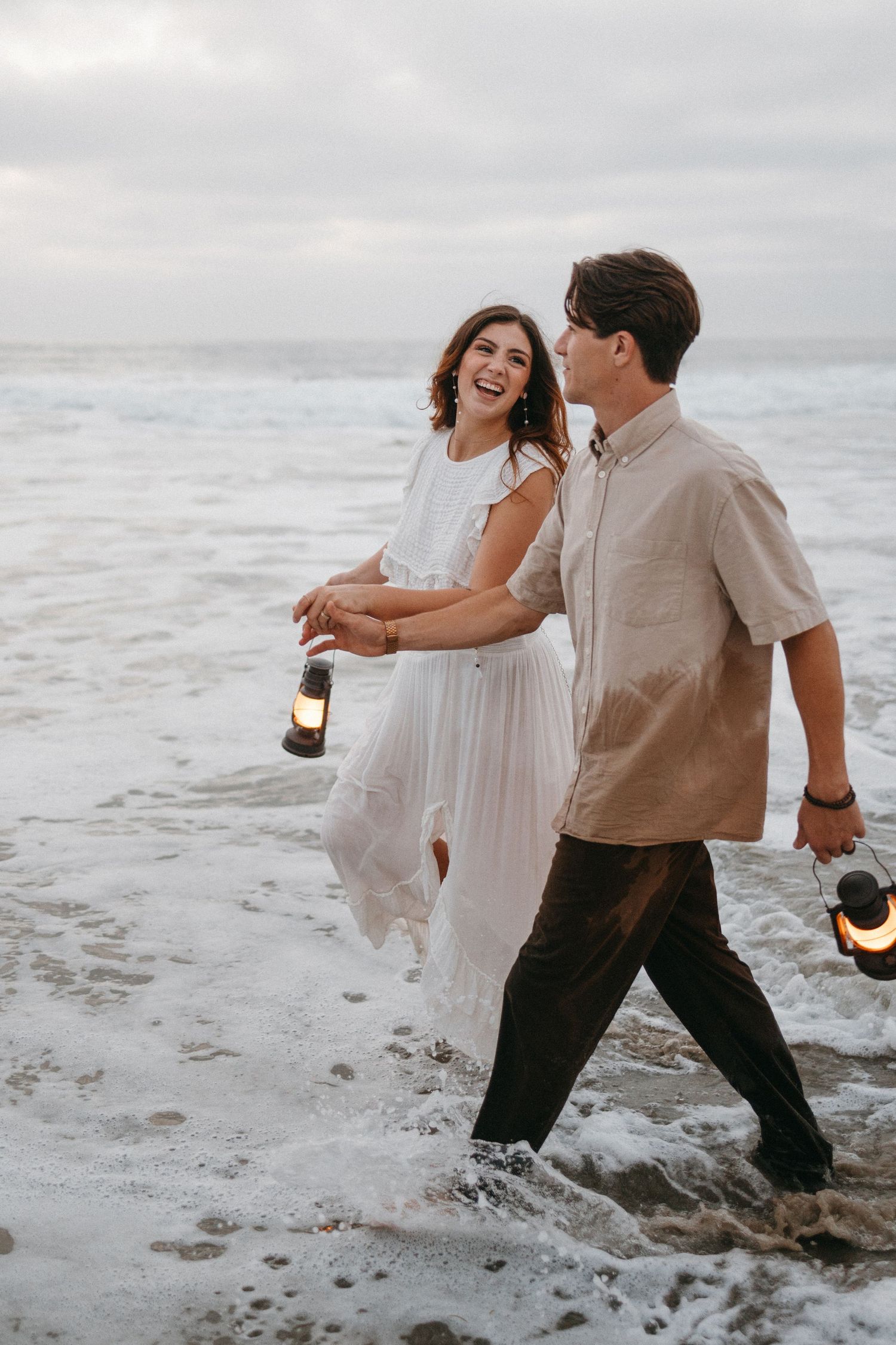 A couple wades through ocean waves at sunset while holding lanterns.