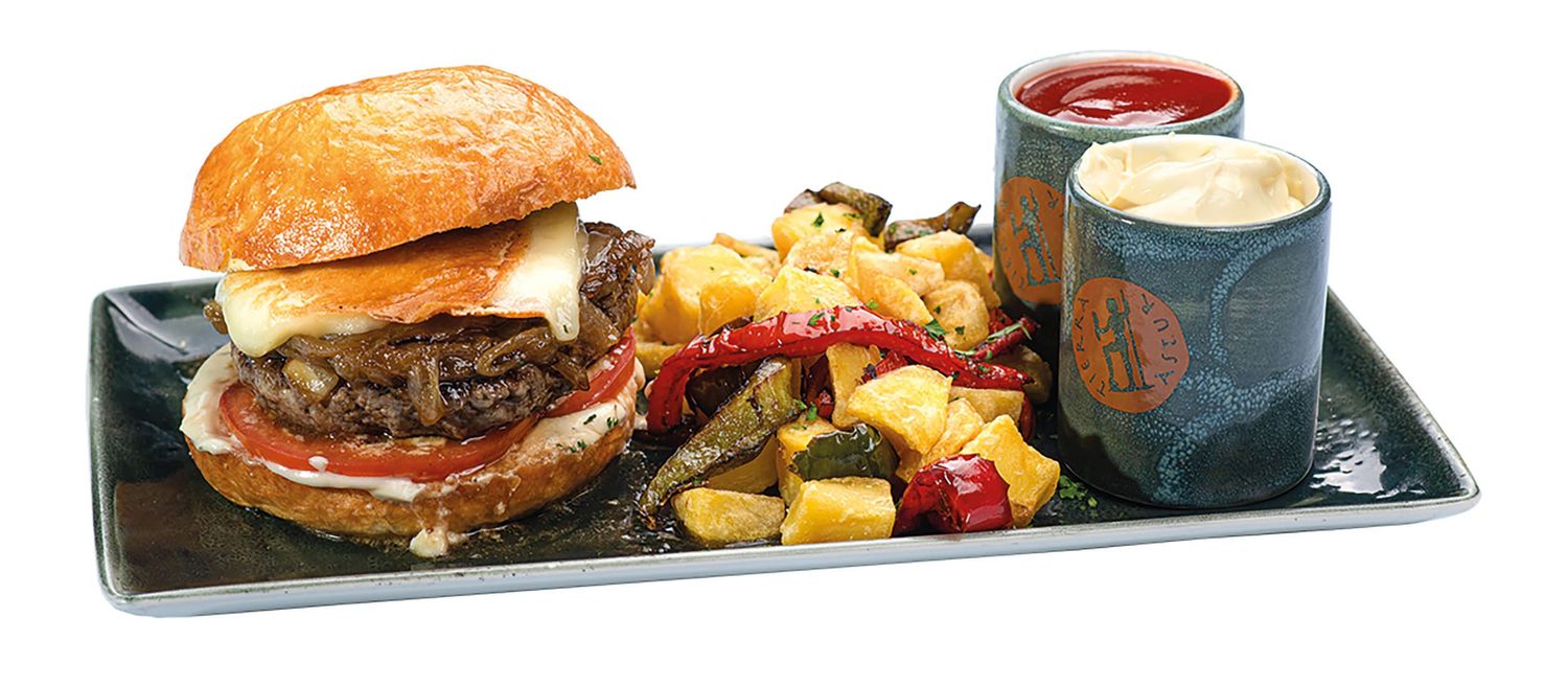 Una hamburguesa gourmet servida en un plato negro con gajos de papa sazonados, ensalada de col y dos salsas para acompañar. Tierra Astur
